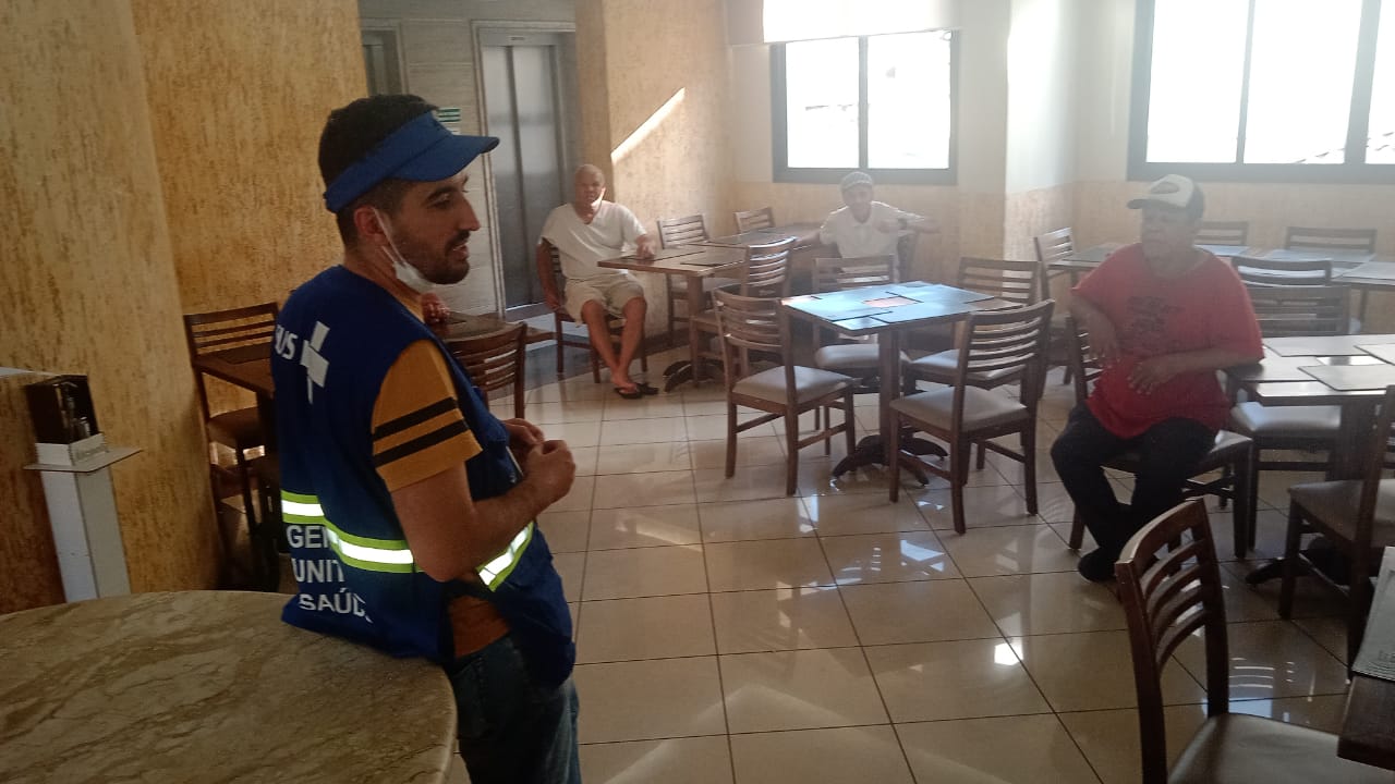 Na imagem, um agente do 'Consultório na rua', ministra a oficina em uma sala com mesas e cadeiras. Podemos ver na imagem, três senhores assistindo a oficina sentados nessas mesas. A sala possui janelas pretas e paredes amarelas.